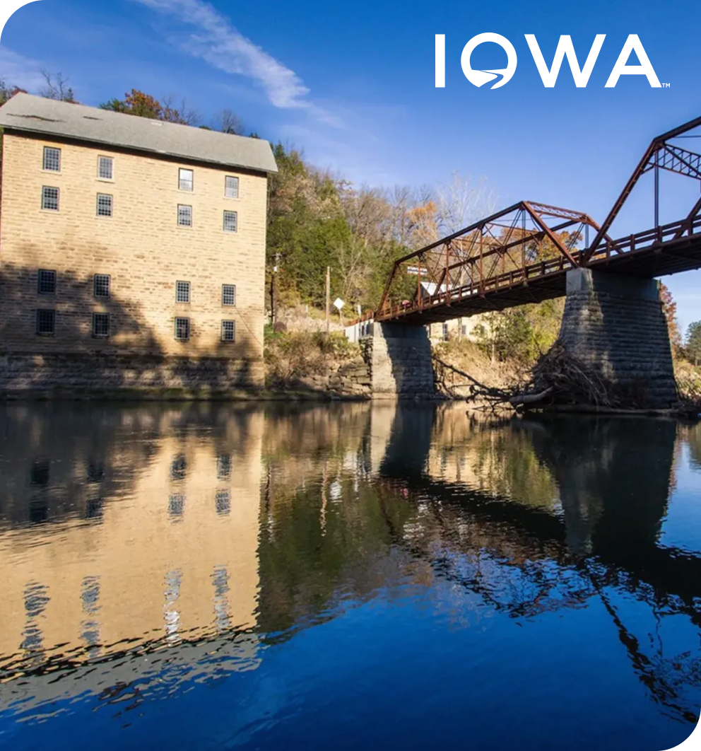 A historic stone mill and iron bridge reflected in a river beneath a blue sky, representing the University of Iowa's use of Qualtrics for employee experience.